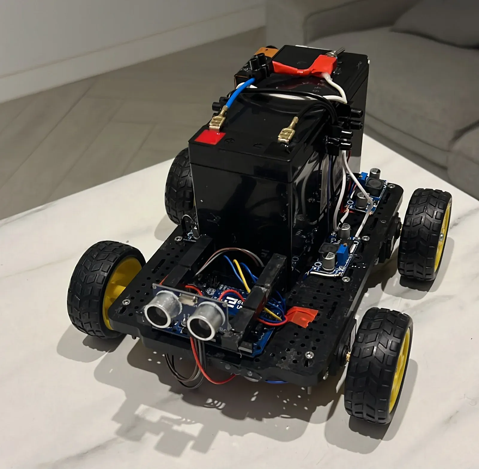 Four-wheeled sumo robot with a black chassis, exposed wiring, batteries, and front-mounted ultrasonic sensors.